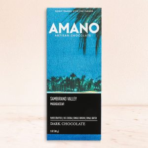 Blue and black chocolate bar packaging with 'Amano Artisan Chocolate' in white. Tropical trees silhouette and text highlight origin: Sambirano Valley, Madagascar.
