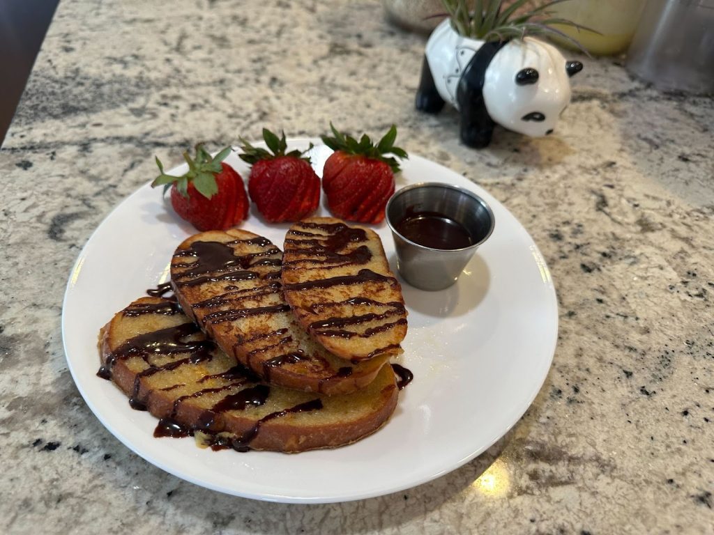 A plate of French toast drizzled with chocolate sauce, garnished with strawberries, and a side of chocolate dip, with a panda figurine in the background.