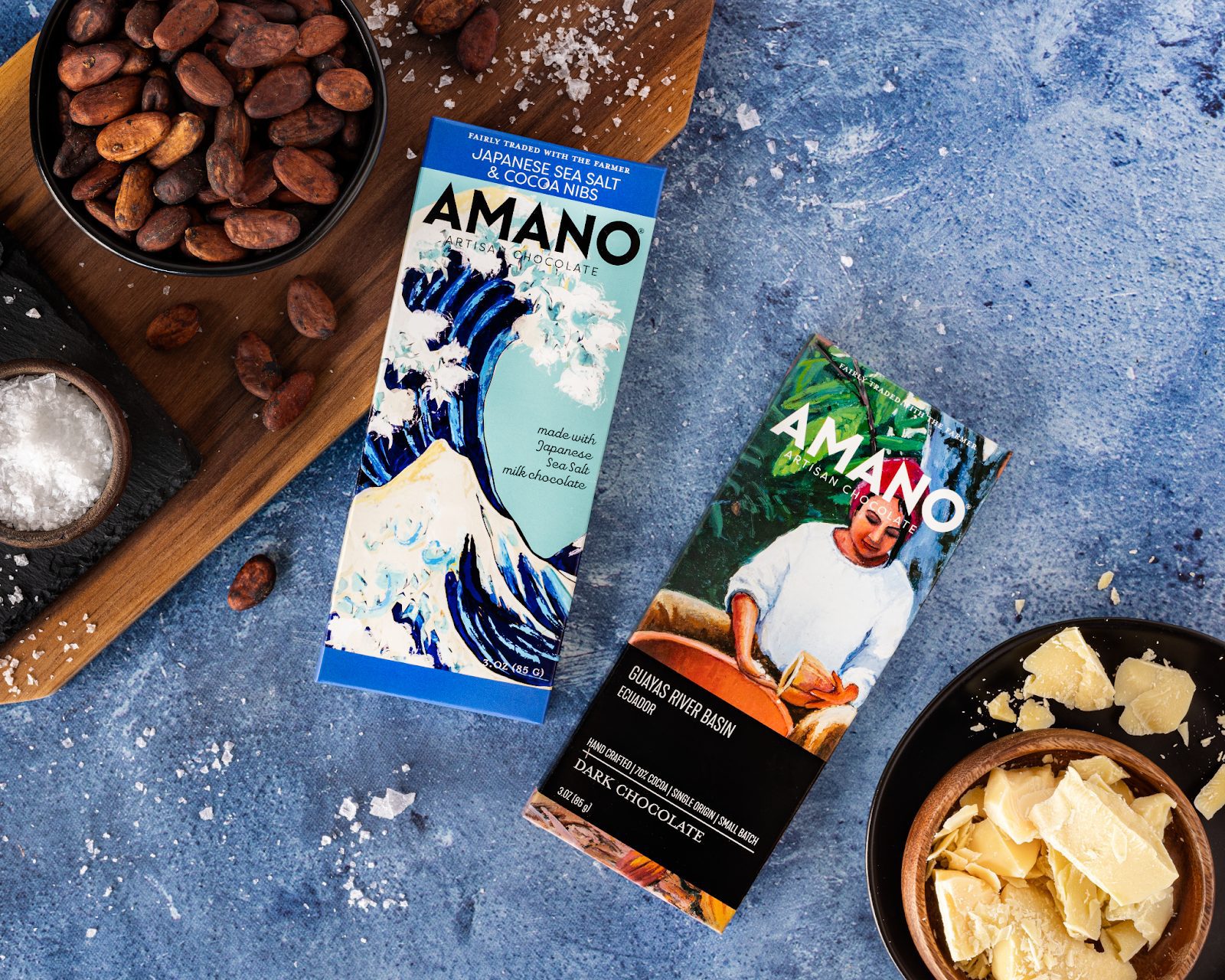 Amano Artisan Chocolate bars with cocoa nibs, sea salt, and dark chocolate, surrounded by cocoa beans and chocolate pieces.