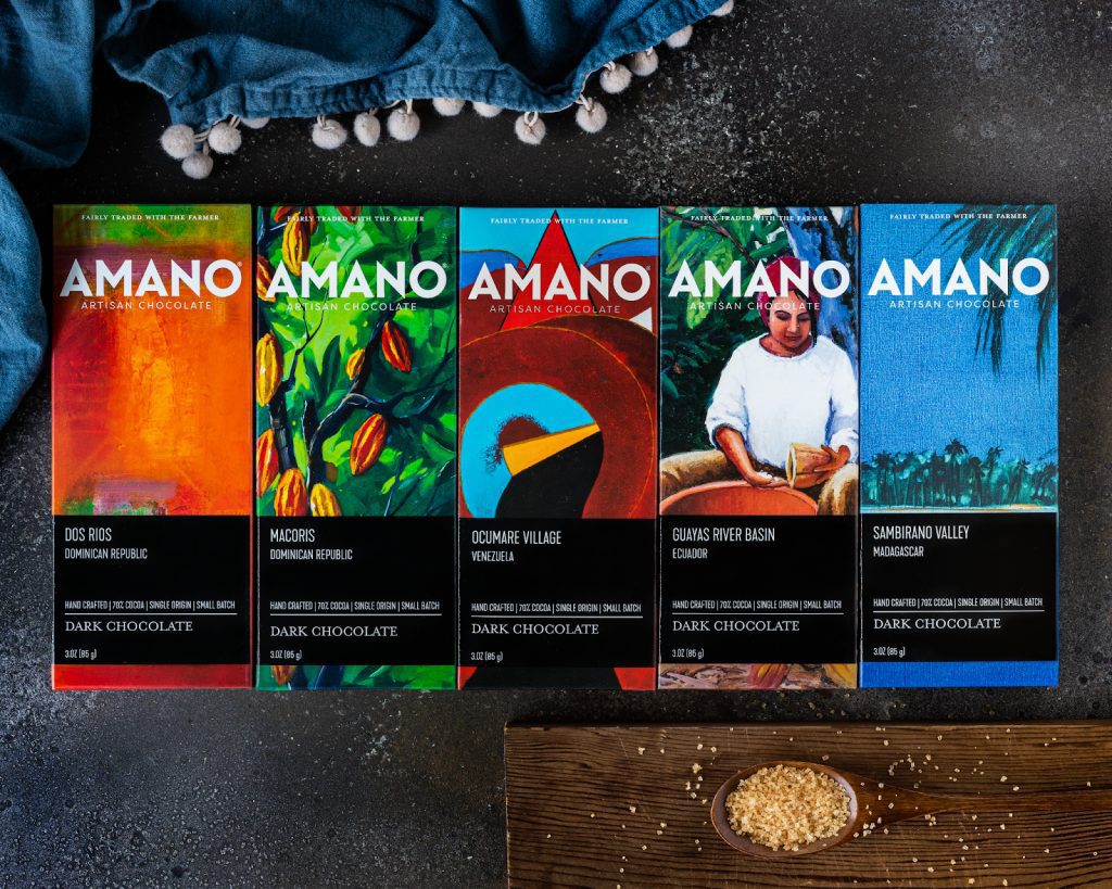Five Amano Artisan Chocolate bars with unique designs and origins, displayed with cocoa beans.