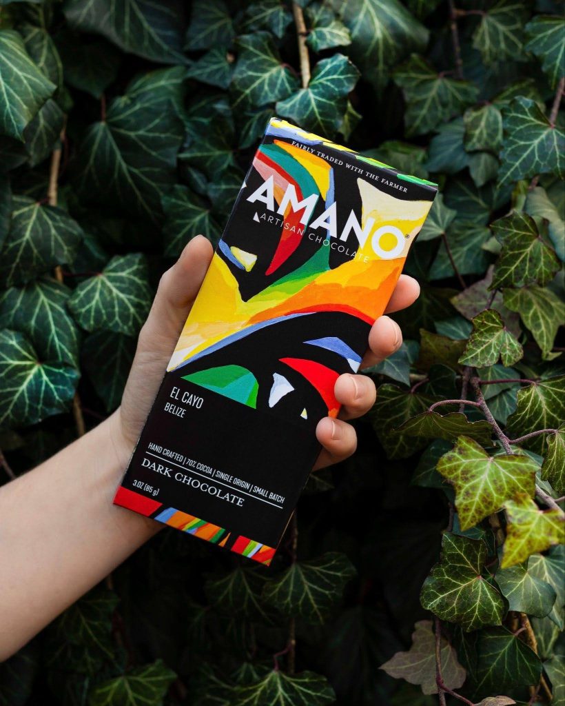 A hand holds a colorful Amano Artisan Chocolate bar against a backdrop of lush green ivy leaves