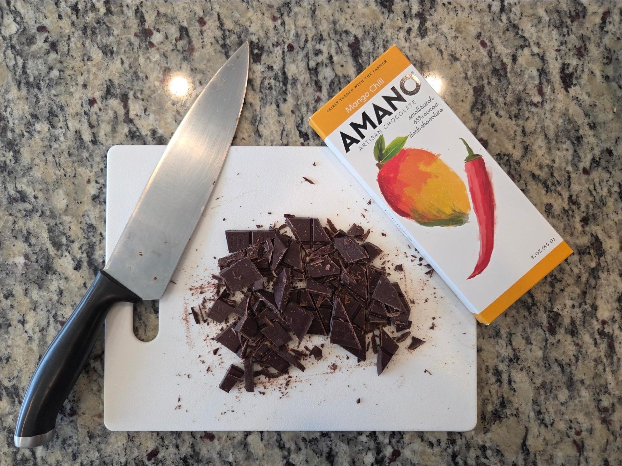Chopped dark chocolate on a cutting board next to a chef's knife and an Amano Mango Chili chocolate wrapper.