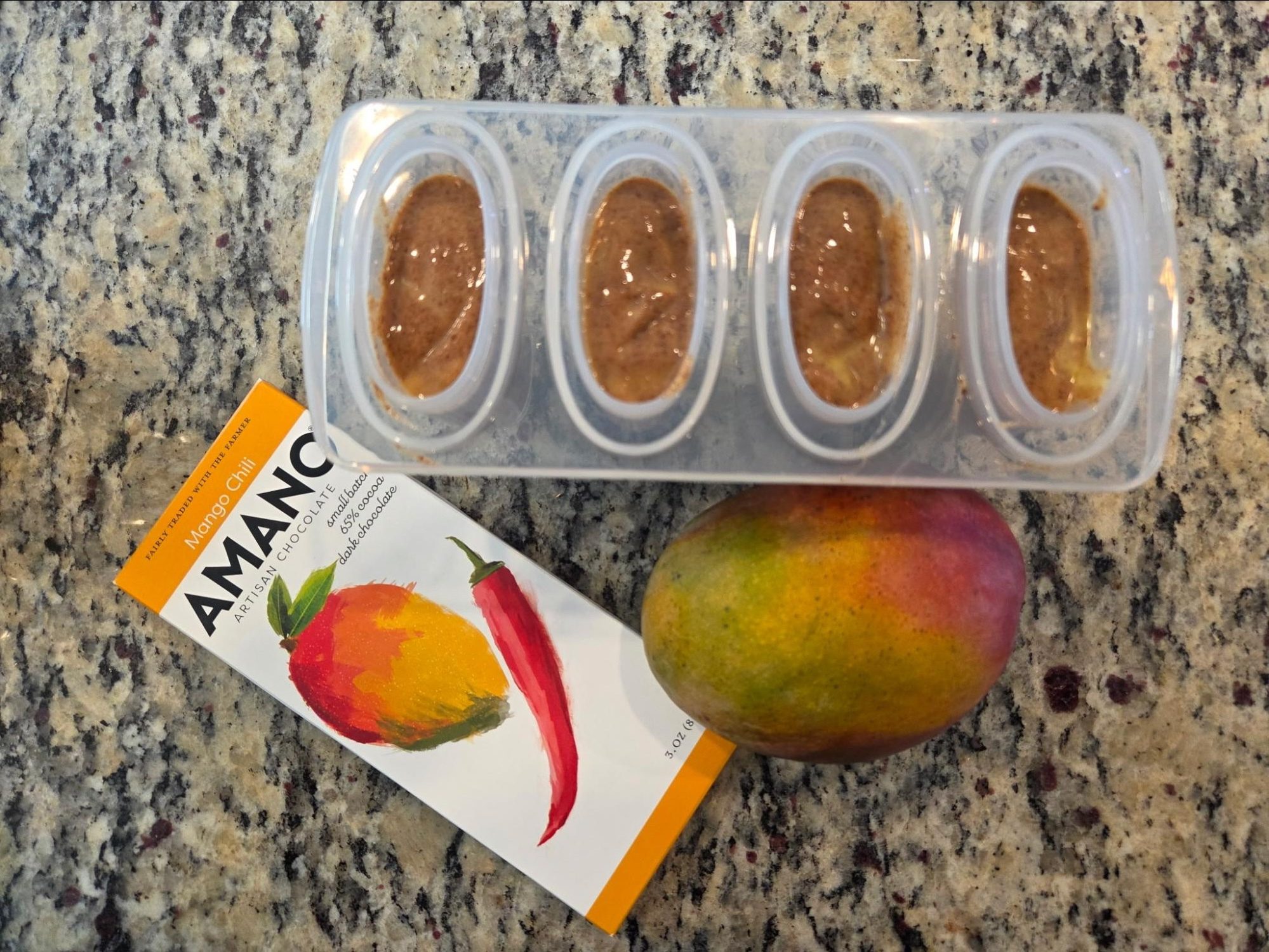 Fresh mango, Amano Mango Chili chocolate bar, and filled popsicle mold on a countertop.