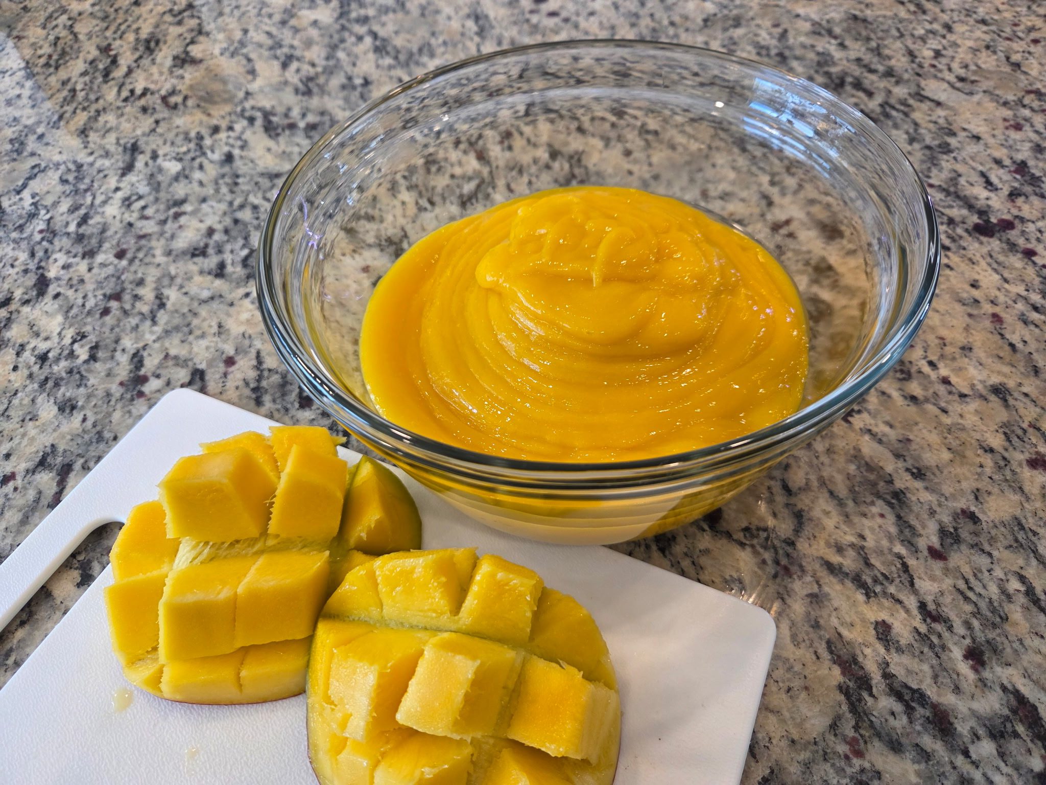 Glass bowl filled with smooth mango puree beside fresh scored mango halves on a cutting board.