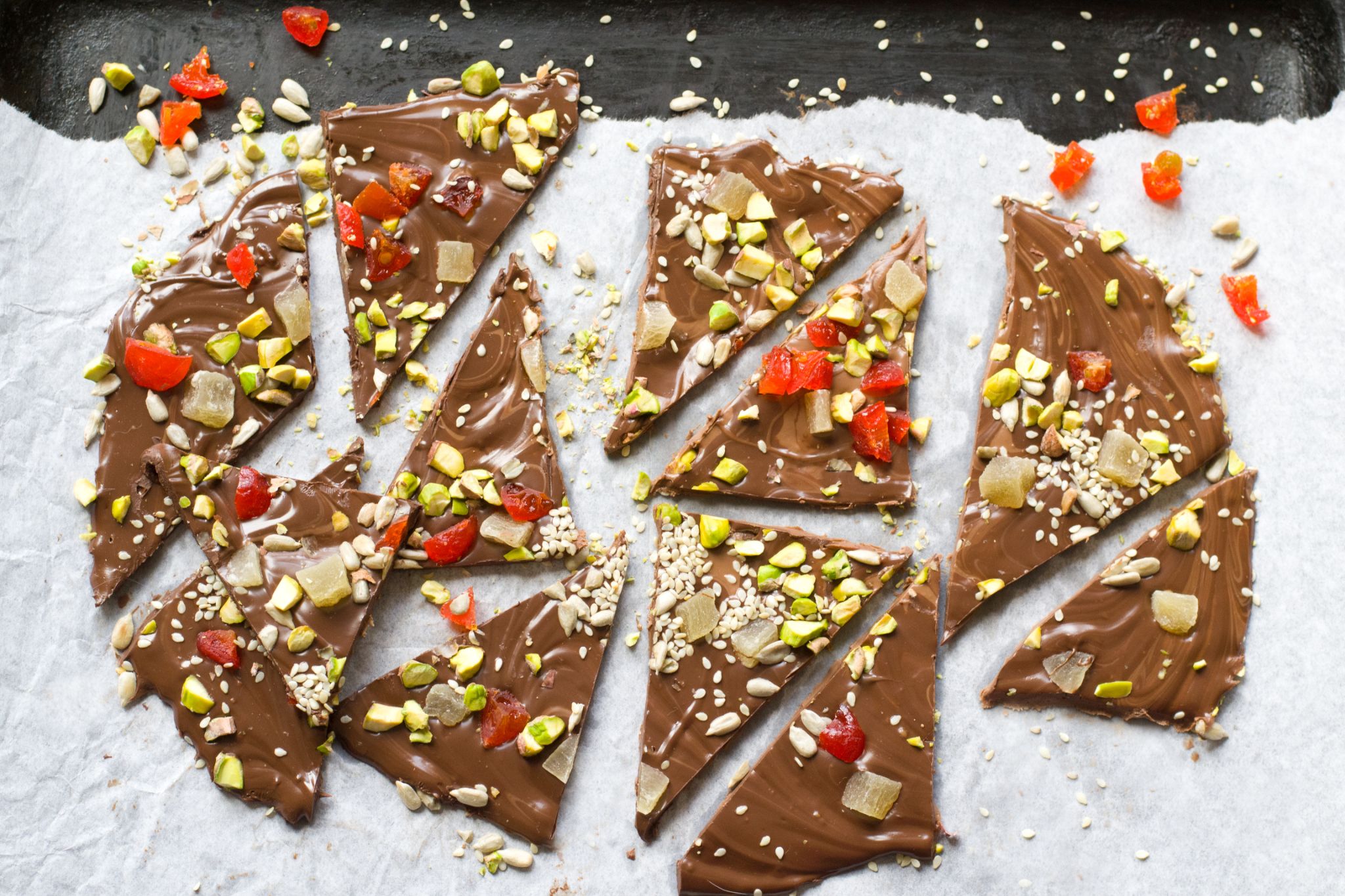 Triangular pieces of milk chocolate bark topped with pistachios, dried fruit, seeds, and sesame seeds on parchment paper.