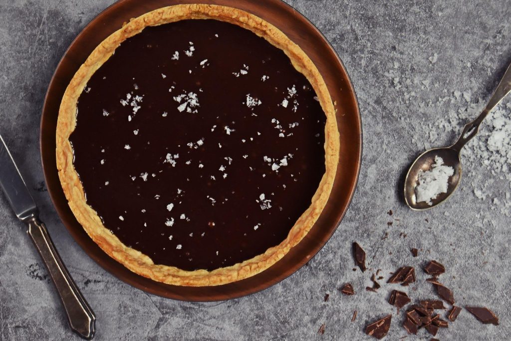 A rustic chocolate tart with a golden crust and scattered white flakes sits on a dark plat